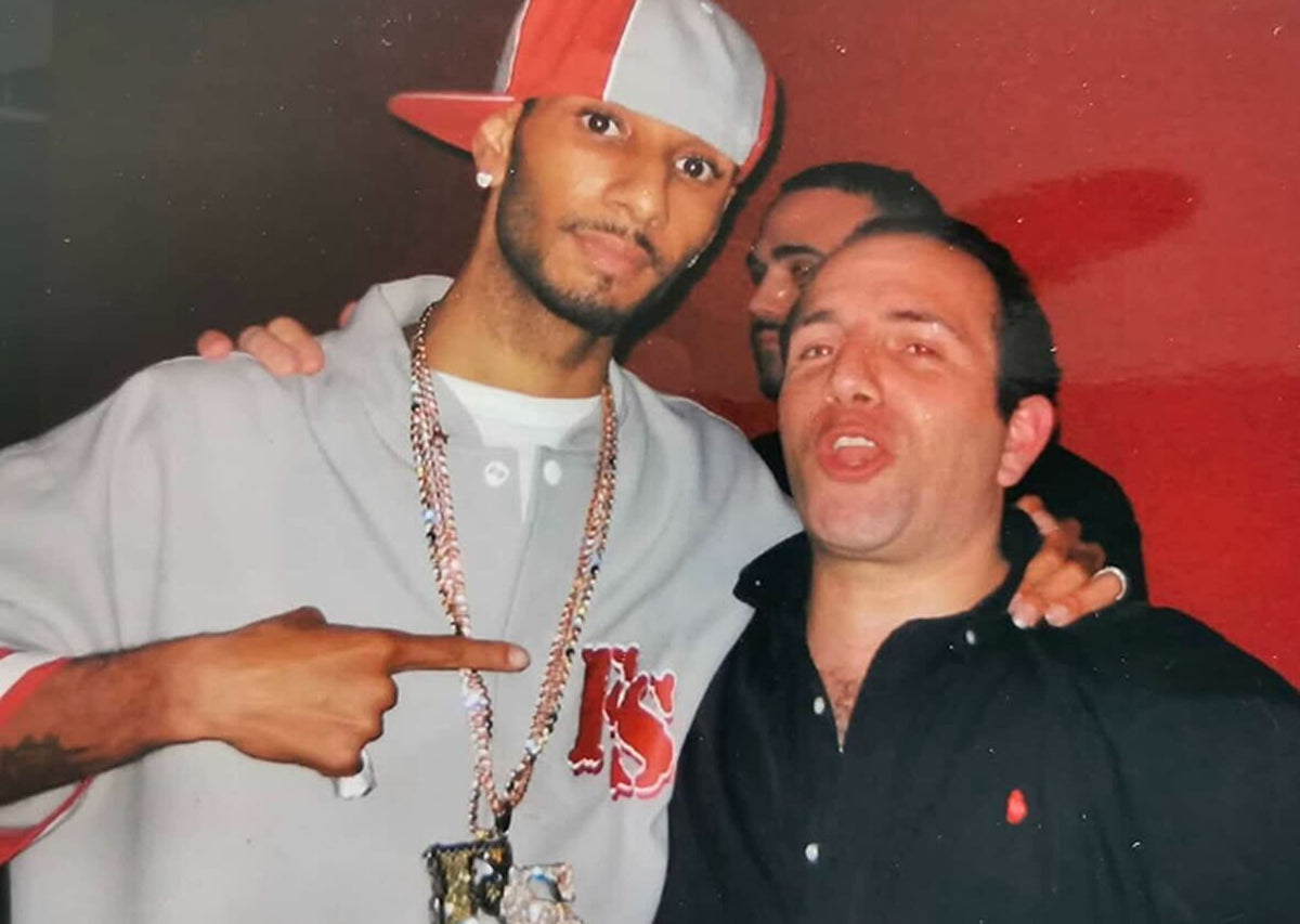 Two men posing together with one pointing, against a red wall.